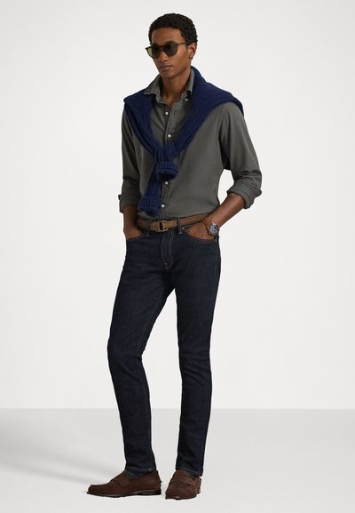 Vīriešu apģērbs ar tumši pelēku pogājamu kreklu, jūras zila adīta džemperīšu, kas novilkts pār pleciem, tumšām džinsām un brūniem mokāniem. Zaļi saulesbrilles.