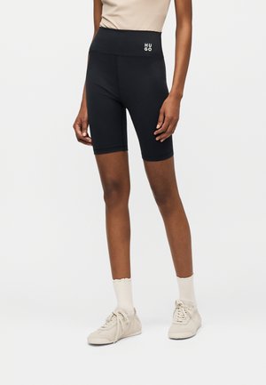 Woman wearing black high-waist biker shorts with "HUGO" logo, white socks, beige sneakers, and a beige sleeveless top on a plain background.