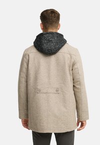 Cappotto beige in misto lana con cappuccio lavorato a maglia scuro, caratterizzato da un taglio dritto, bottoni sul retro e linee pulite. La texture appare morbida e calda.