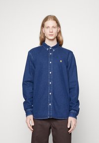 Camisa de denim azul oscuro con un diseño de botones, que cuenta con un cuello y un logo en el pecho. Corte ajustado con mangas largas y puños de botón.