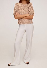 Vaaleanbeige pitsitoppi, jossa on muhkeat hihat, yhdistettynä valkoisiin leveälahkeisiin housuihin. Asussa on kukkakuvio ja sileät tekstuurit.