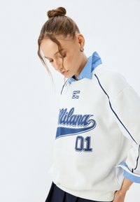 Sudadera blanca con texto gráfico azul "Milana 01" y detalles azules, combinada con una camisa de cuello celeste debajo.