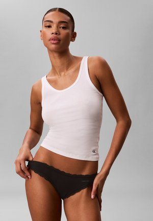 Femme portant un débardeur blanc et des sous-vêtements noirs avec bordure en dentelle, debout devant un fond gris uni.
