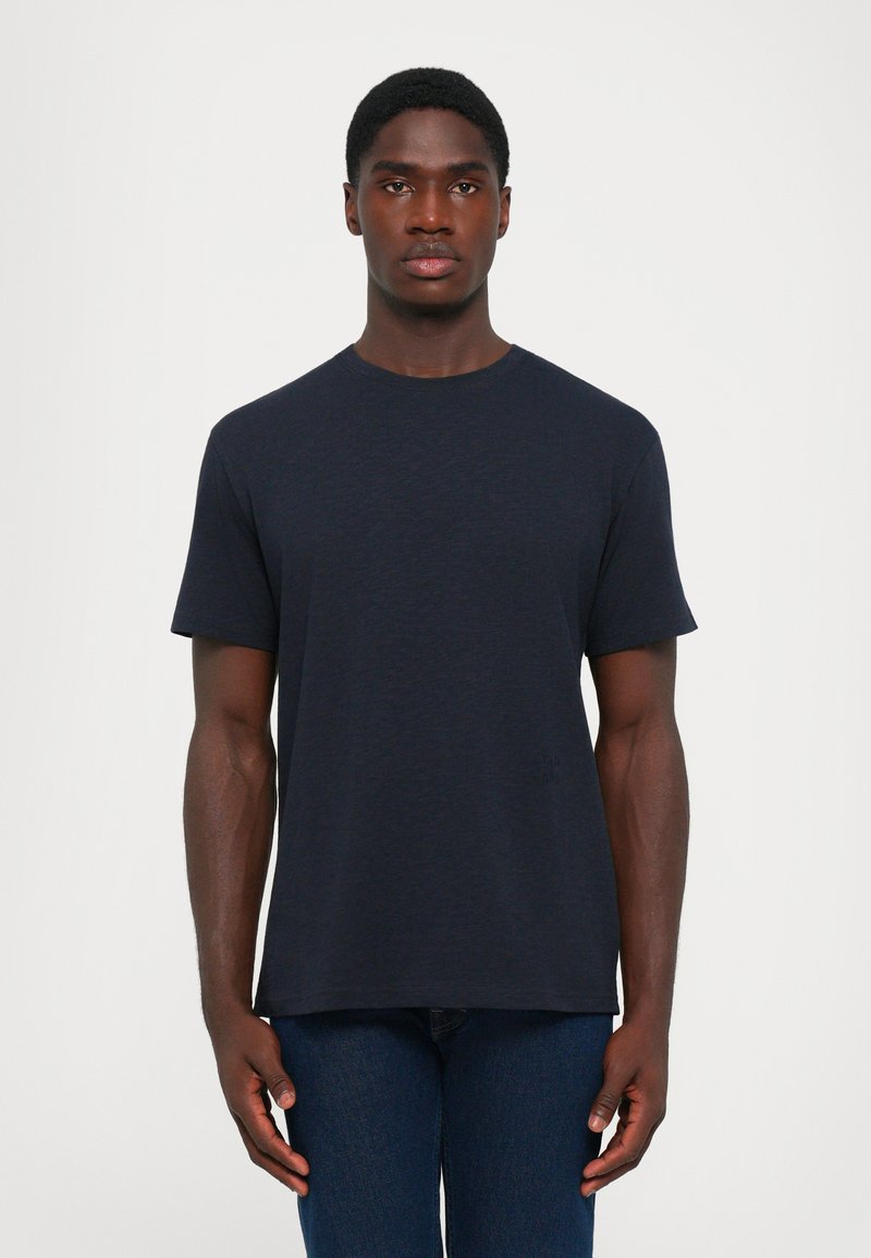 Man wearing a plain dark navy short-sleeve t-shirt and dark blue jeans standing against a white background.