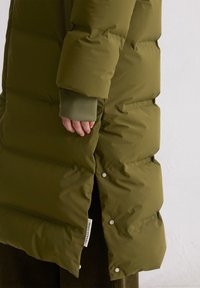 Olive zielona kurtka puchowa z pikowanymi segmentami, bocznymi zapięciami na zatrzaski i ukrytymi mankietami. Zawiera metkę tkaninową w pobliżu dolnej krawędzi.