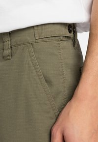 Pantalones verde oliva con un tejido texturizado, que cuentan con un cierre de botón, presillas para el cinturón y bolsillos laterales. Acentos cosidos visibles.