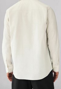 Camisa de manga larga en tejido beige claro, con un dobladillo curvado y puños abotonados, que presenta un diseño liso y texturizado en la parte trasera.