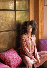 Long-sleeved pink floral top paired with striped pants, sitting on a wooden bench with vibrant patterned pillows in a warm indoor setting.