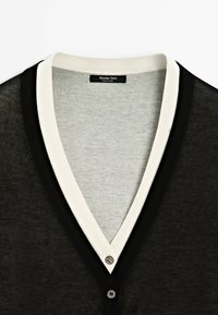 Cardigan noir avec bordure blanche et deux boutons noirs au col en V, montrant une doublure intérieure grise et une étiquette Massimo Dutti.