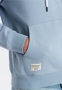 Ljusblå hoodie i bomullsblandning, med en framficka, ribbade mudd, och en etikett på sidan med text och logotyp.