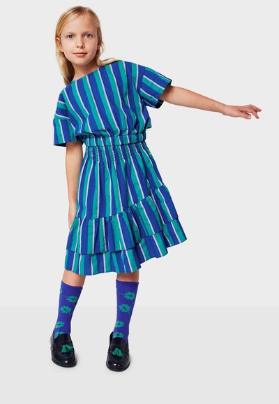 Abito a righe blu e verdi con vita arricciata e orlo arricciato, abbinato a calzini viola con motivi floreali e scarpe nere.