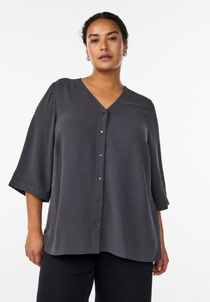 Žena nosi voľnú tmavosivú blúzku na gombíky s tromi štvrťovými rukávmi a čierne nohavice, na jednoduchom bielom pozadí.