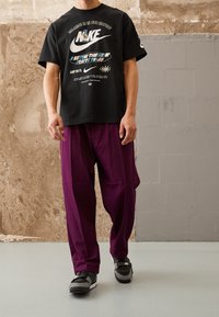 Černé bavlněné tričko s grafickým potiskem a logem Nike, doplněné volnými tmavě fialovými kalhotami a černými teniskami s logem.