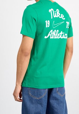 Osoba nosząca zieloną koszulkę Nike z białym napisem "Nike Athletics 1972" i logo Swoosh na plecach, w połączeniu z niebieskimi szortami z denimu.
