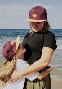Casquette bordeaux avec un bord jaune et un logo, portée par une femme et une fille sur une plage. Les deux sont en vêtements décontractés ; une interaction ludique est visible.