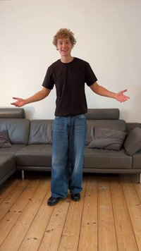 Camiseta negra de algodón, jeans anchos de pierna en azul claro y zapatos negros usados por una persona que está de pie sobre un suelo de madera en una sala de estar con un sofá gris.