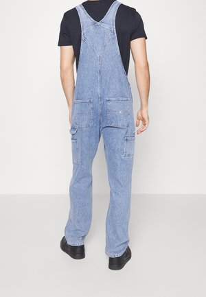 Personne portant une salopette en denim bleu clair sur un t-shirt noir, debout, dos tourné vers la caméra sur un fond uni.