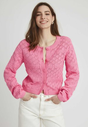 Cardigan en tricot rose avec une texture en dentelle, présentant un col rond, des manches longues et une fermeture à boutons à l'avant. Associé à un pantalon blanc.