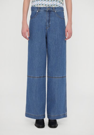 Jean en denim bleu à large jambe avec une ligne d'ourlet cousue, taille haute et deux poches avant, associé à un haut à motifs et des chaussures noires.