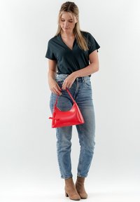 Bolso de cuero rojo con forma curva, asa corta y un lazo decorativo, combinado con una blusa oscura y jeans azules.
