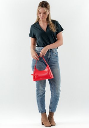 Sac à main en cuir rouge avec une forme arrondie, une courte poignée et un accent de nœud décoratif, associé à un haut sombre et un jean bleu.