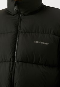 Sort puffersjakke med høj krave, der har en frontlynlås og quiltet polstring. Carhartt-logo i grå på brystområdet.