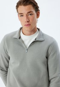 Maglione grigio chiaro con zip, caratterizzato da una finitura texture, con colletto alto e zip a contrasto. Indossato sopra un sottogiacca bianco.