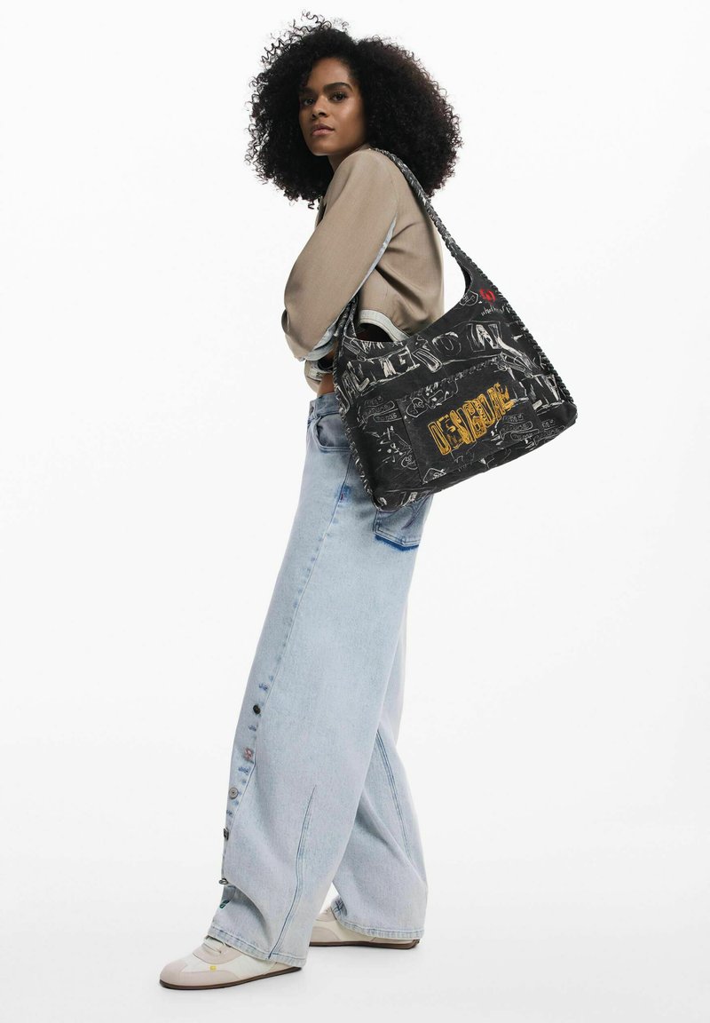 Must kandesuurusega õlakott graafilise trükiga, mis kujutab visandeid ja kollase värviga sõna "DESIGNA". Pehme materjal, avar disain ja kootud detailid.