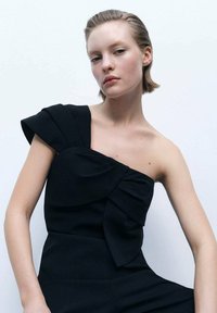 Black one-shoulder jumpsuit with a textured bow detail on the chest, featuring a fitted design and smooth fabric.