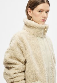 Beežist fliisist jak, millel on kõrge krae, kuldne tõmblukk ja tekstureeritud kangas, sile eestmine paneel. Märkimisväärsed liialdatud varrukad ja taskud.