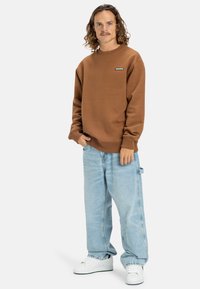 Sudadera marrón de cuello redondo con puños y dobladillo acanalados, combinada con jeans holgados de color azul claro. Zapatillas blancas completan el atuendo.