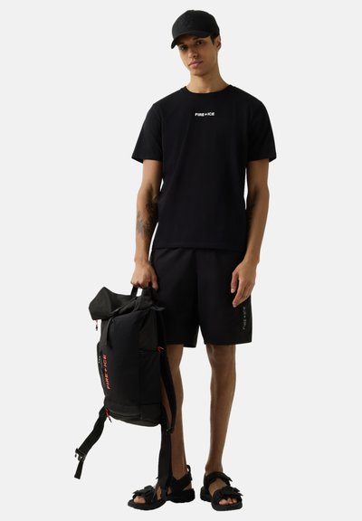 Jeune homme portant une casquette noire, un t-shirt noir "FIRE+ICE" et un short, tenant un sac à dos noir, debout devant un fond blanc.