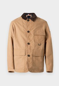 Brun jakke lavet af bomuld, med en kordfløjlskrage, fem knapper, to forlommer og en metalring som detalje. Glat tekstur.