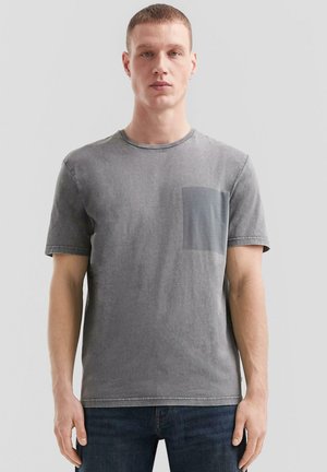 Junger Mann mit kurzen Haaren, der ein ausgewaschenes graues T-Shirt mit kurzen Ärmeln und Brusttasche sowie dunkelblaue Jeans trägt, steht vor schlichtem Hintergrund.