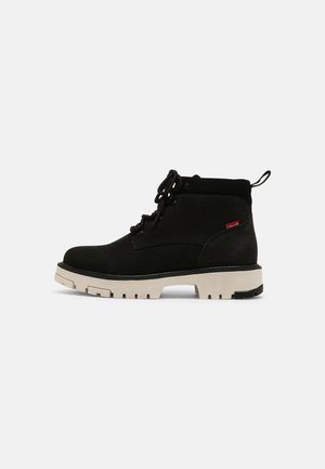 Black suede ankle boots with a round toe, beige chunky sole, matching laces, and a short cuff. Features a red logo tag on the side.