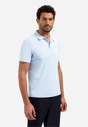 Man met donker krullend haar en baard, gekleed in een lichtblauw poloshirt met korte mouwen en een donkere broek, staand tegen een effen achtergrond.