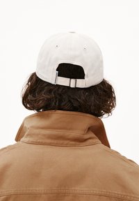 Beige katoenen pet met verstelbare band aan de achterkant, gedragen door een persoon in een bruine jas met kraag. Achteraanzicht toont het gestructureerde materiaal.