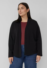 Vrouw draagt een losse zwarte cardigan met open voorkant over een donkerrode top en blauwe spijkerbroek, staand tegen een effen lichtgrijze achtergrond.