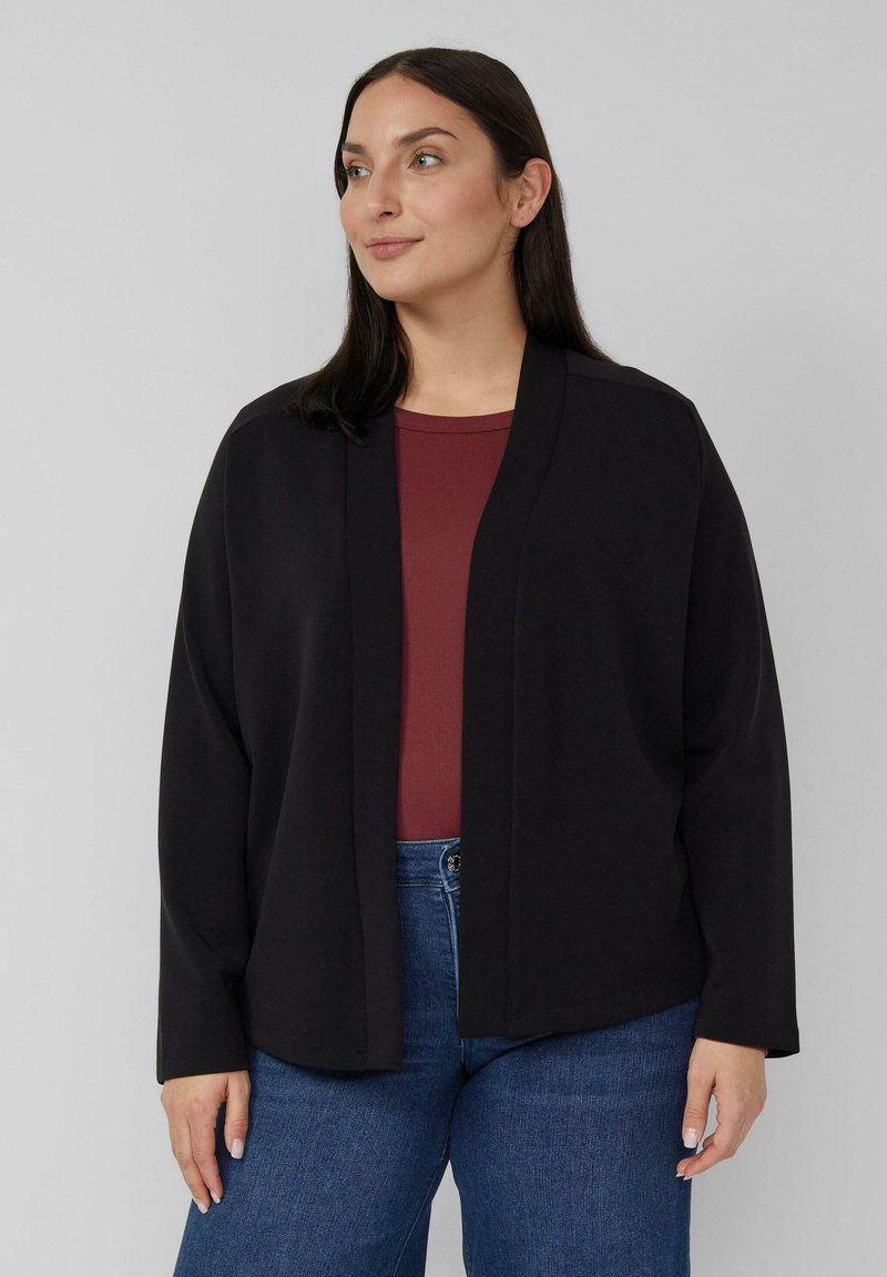 Vrouw draagt een losse zwarte cardigan met open voorkant over een donkerrode top en blauwe spijkerbroek, staand tegen een effen lichtgrijze achtergrond.