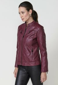 Veste en cuir bourgogne avec col mandarin, poches zippées et détails matelassés aux épaules, associée à un pantalon noir texturé.