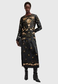 Rochie neagră din paiete cu mâneci lungi, prezentând broderie aurie cu stele și păsări, și fante laterale. Asortată cu cizme negre până la genunchi.