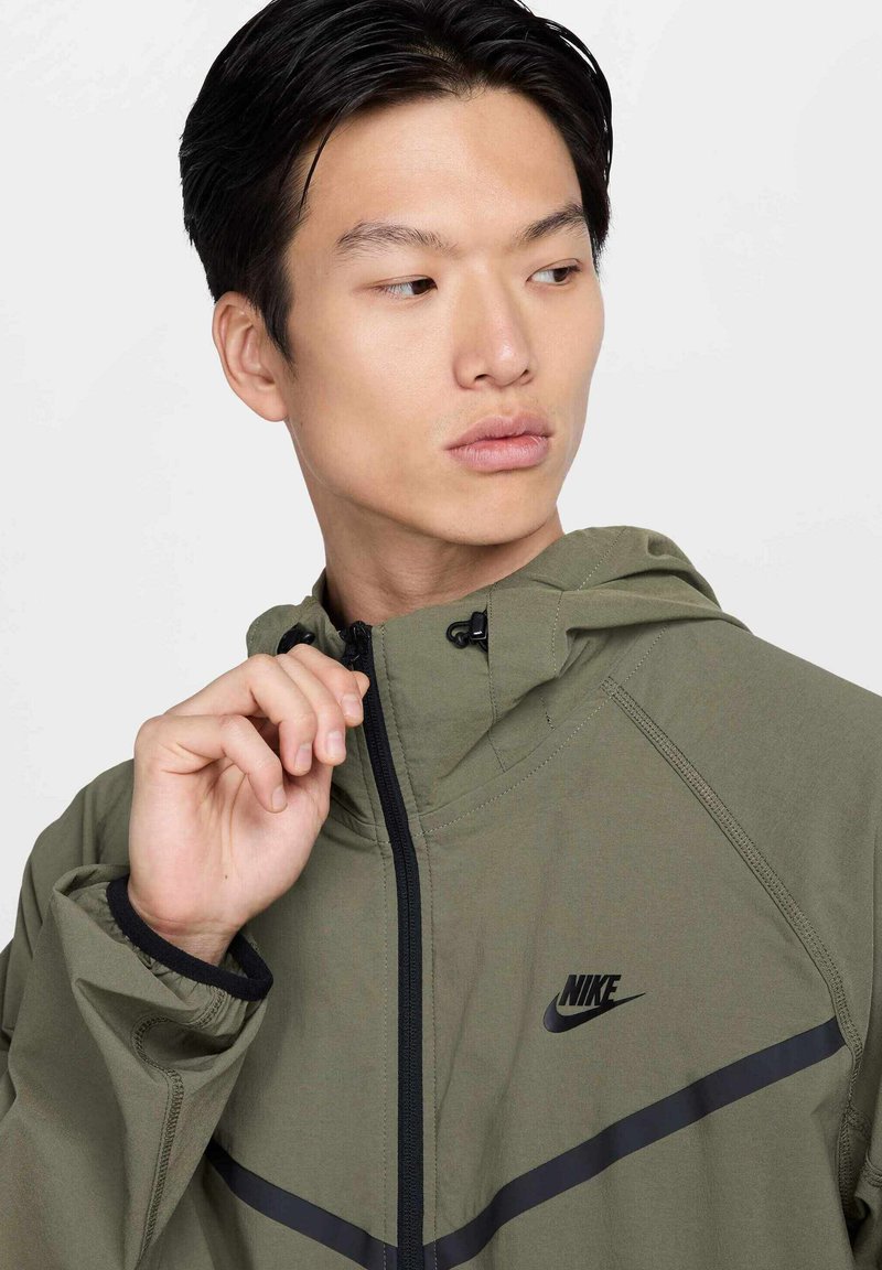 Oliwkowa kurtka Nike z kapturem, z czarnym zamkiem i czarnymi paskami akcentującymi na klatce piersiowej. Gładka faktura i elegancki design.