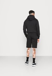 Felpa nera con tasca frontale, abbinata a pantaloni corti neri. L'uomo indossa calzini neri e sneakers bianco e nere. Variazioni della pelle visibili.