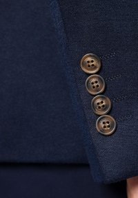 Morski żakiet z rękawem ozdobionym czterema okrągłymi brązowymi guzikami. Każdy guzik ma dwa otwory i wygrawerowane logo. Gładka, teksturowana tkanina.