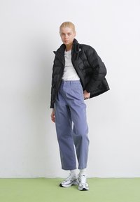 Schwarze Pufferjacke, weißes T-Shirt, lavendelfarbene Hose und silberne Sneaker. Strukturiertet Stoff und lockere Passform in einem neutralen Umfeld.