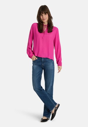 Frau mit langen braunen Haaren, die eine leuchtend pinke Langarmbluse, blaue Jeans und schwarze spitze Ballerinas trägt, steht mit gekreuzten Beinen.