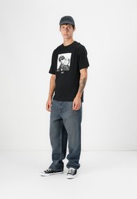 Black short-sleeve t-shirt with a photographic print on the chest, paired with loose blue denim jeans and black sneakers.