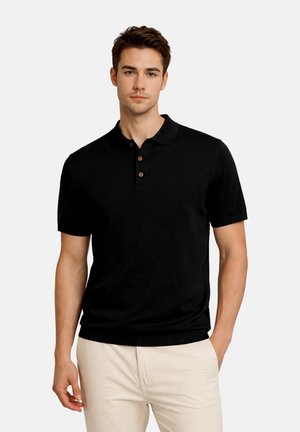 Junger Mann, der ein schwarzes Poloshirt mit kurzen Ärmeln und drei Knöpfen sowie hellbeige Hosen trägt, steht mit einer Hand in der Tasche.