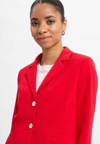 Roter Blazer mit Reverskragen, zwei Knöpfen und seitlichen Taschen. Glatte Stofftextur mit weißen Knopfakzenten. Über einem hellen Hemd getragen.