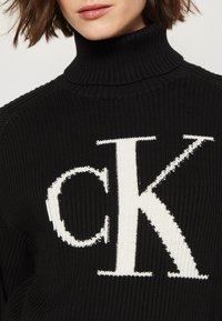 Maglione nero a collo alto in maglia, caratterizzato da un grande logo bianco "CK" sul davanti. La texture è a coste e il colletto è alto.